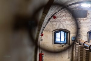 Interior view of a historical building showing a brick wall with arched windows, a red fire extinguisher, metal piping, and an out-of-focus object in the foreground.