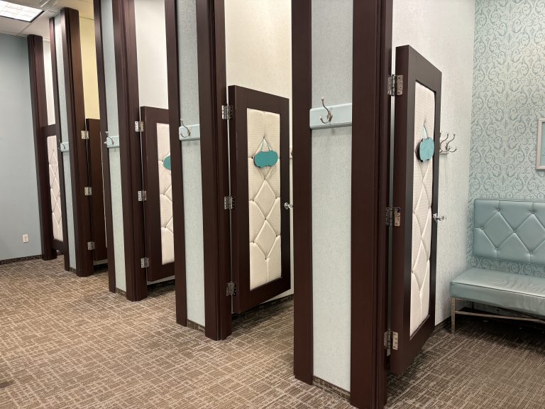 A row of fitting rooms in a clothing store with open doors each featuring a decorative quilted pattern. Hooks are next to each door on the outside wall.  A light blue bench sits against the wall with patterned wallpaper on the right.