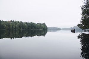 A serene lake with a mirror-like reflection, showing trees and hill silhouettes in a foggy atmosphere.
