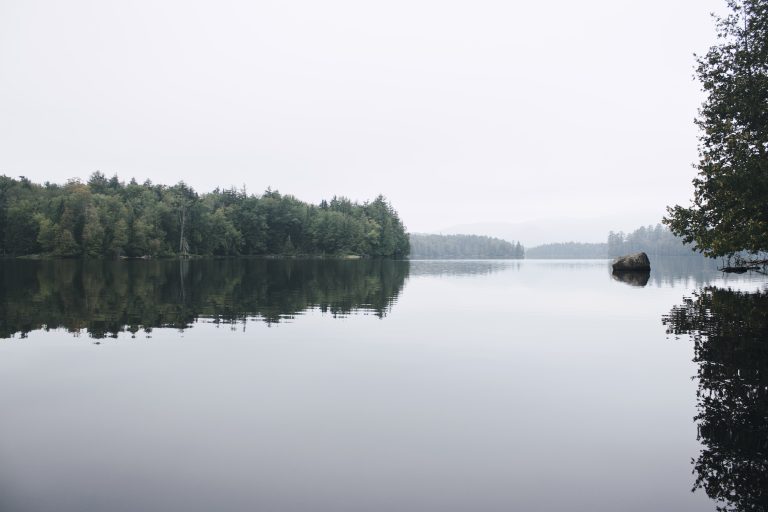 A serene lake with a mirror-like reflection, showing trees and hill silhouettes in a foggy atmosphere.