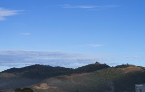 A landscape view in Cartago, Costa Rica featuring rolling green hills under a blue sky with wispy clouds. There’s a visible smoke plume near the bottom right corner, indicating human activity, and the corner of a building can be partially seen on the right edge of the frame.
