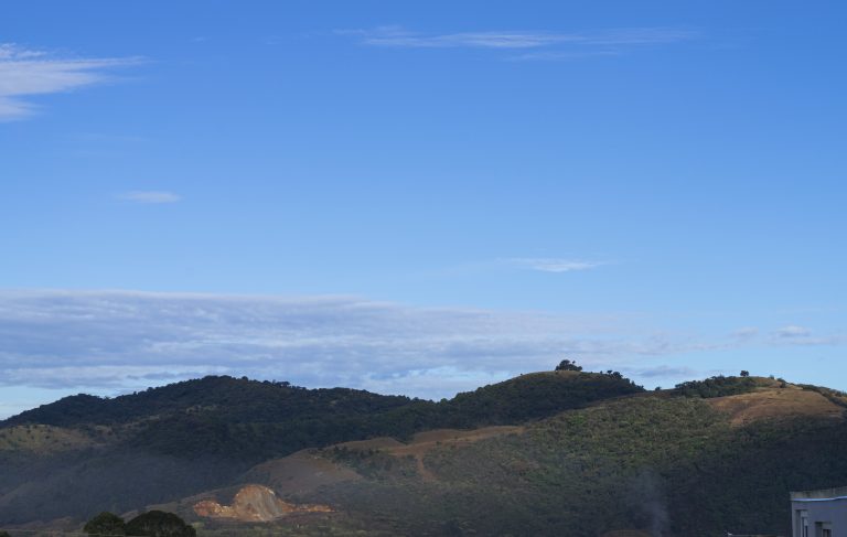 A landscape view in Cartago, Costa Rica featuring rolling green hills under a blue sky with wispy clouds. There’s a visible smoke plume near the bottom right corner, indicating human activity, and the corner of a building can be partially seen on the right edge of the frame.