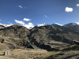 The barren mountainous landscape of Damodar Kunda Upper Mustang under a clear blue sky with some snow-capped peaks in the distance and a small structure and a horse in the foreground.