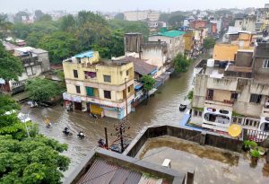Kolhapur, Maharashtra, India experiencing flooding in 2021 with several buildings partially submerged in water and people navigating the flooded streets on motorcycles and by walking.