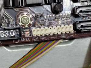 Close-up of a motherboard’s front panel connectors with labels for power LED, speaker, reset switch and power switch, alongside a multi-colored ribbon cable partially connected to the pins.
