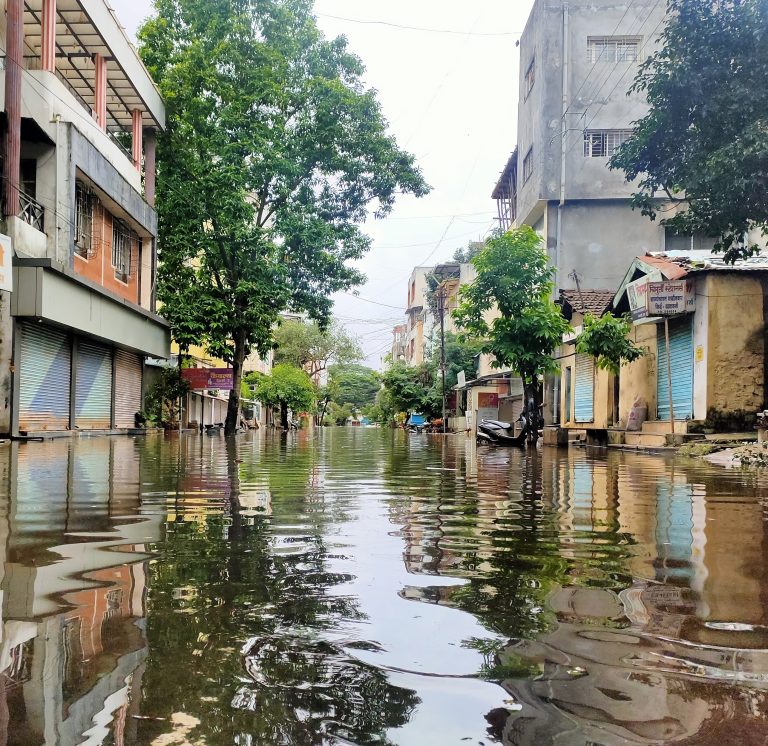 A flooded street with closed shops and trees reflecting in the water, indicating recent flood event in Kolhapur, Maharashtra, India.