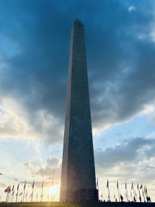 华盛顿纪念碑是位于华盛顿特区国家广场上的方尖碑，落日的余晖照亮了它的背光，天空布满云朵，周围环绕着一圈美国国旗。
