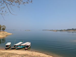 Four colorful wooden boats moored on the shore of Kaptai Lake, Bangladesh with gentle ripples on the water's surface. A clear blue sky above and a quiet, natural landscape with trees and grassy banks in the background.