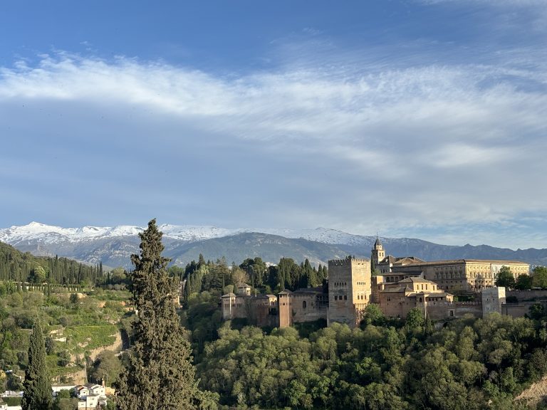 A panoramic view of the Alhambra in Granada., a palatial fortress complex with a backdrop of the Sierra Nevada mountains covered in snow, under a blue sky with scattered clouds.