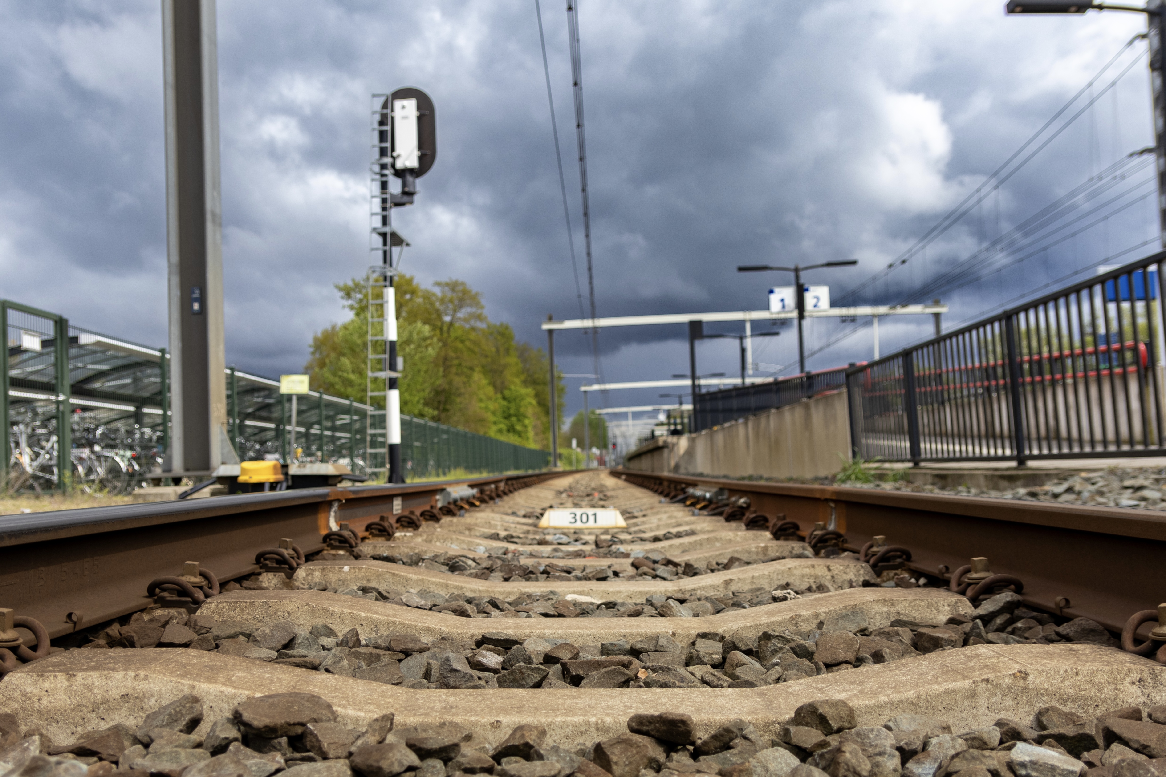 A ground-level view of train tracks leading into the distance with a railway signal on the left, platform numbers in the background, and stormy skies overhead.