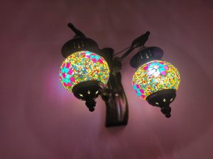 A colorful Turkish mosaic lamp with twin globes hanging from a black metal fixture against a mauve wall, illuminated to display a vibrant pattern of light.

