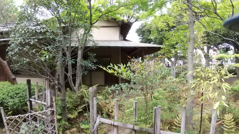 A traditional Japanese tea house surrounded by lush greenery and bamboo fencing, with various plants and trees contributing to a serene atmosphere.