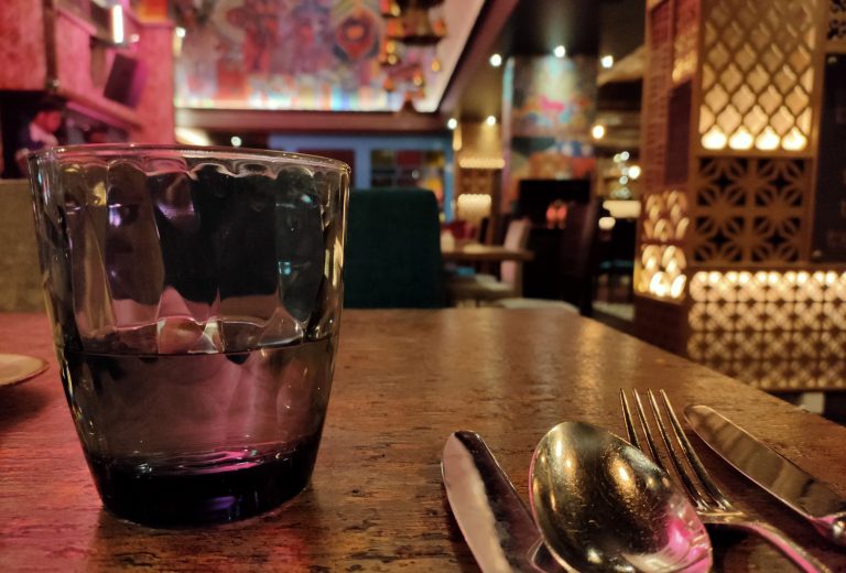 Close-up of a glass with a spoon, fork, and knife on a wooden table with an out-of-focus restaurant interior in the background.