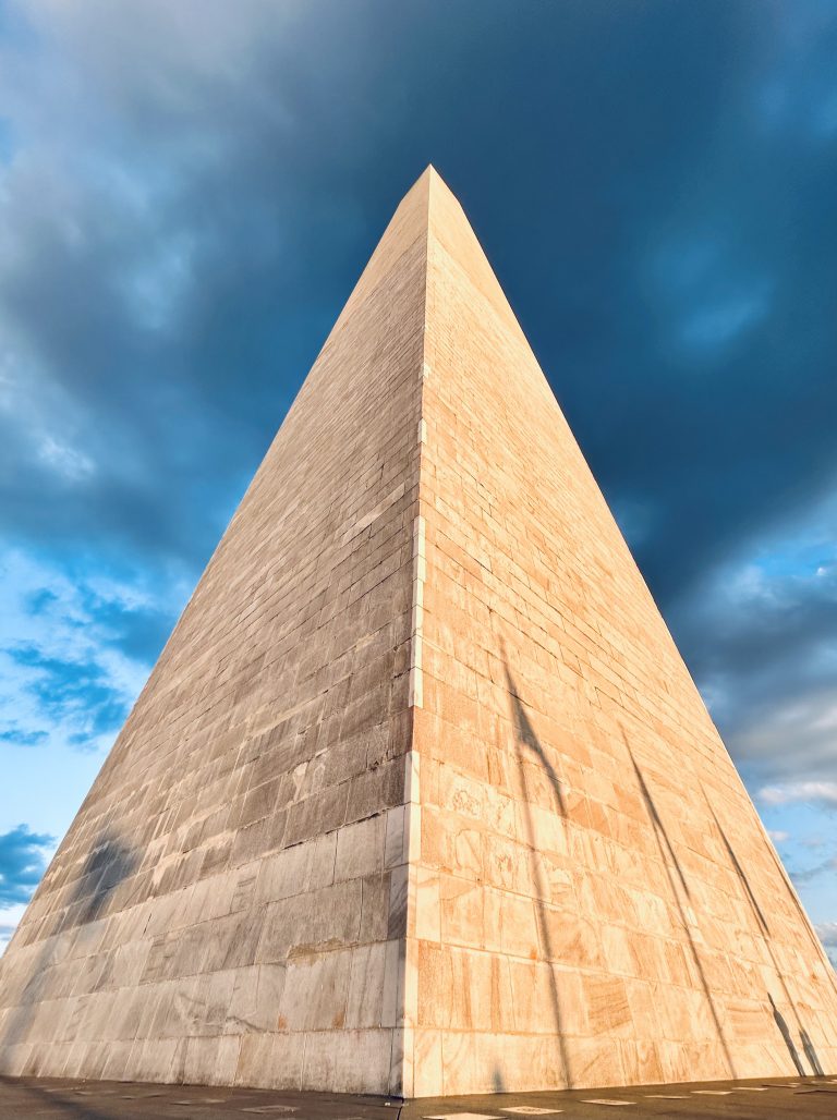 An upward view of the Washington Monument, highlighting its towering height and the contrast of its stone against a bright blue sky with wisps of white clouds.