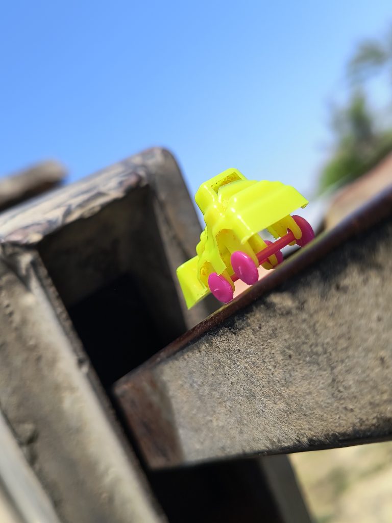 Close-up of a yellow toy car (Bulldozer) with pink wheels on a rusty metal surface (Hand Pump), with a clear blue sky in the background.