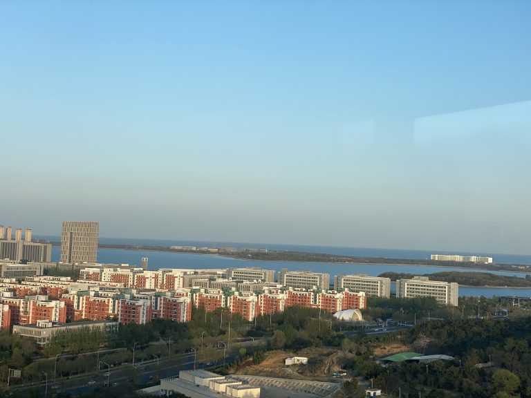 A panoramic view from a high vantage point showing a cityscape with a cluster of red and white residential buildings, a large body of water in the distance, and clear blue sky above during daylight.