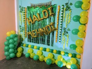 Decorated setup for a Mehandi and Haldi occasion, featuring green and yellow balloons, tinsel curtain backdrop, and golden letter balloons spelling out 'HALDI & MEHNDI'. The vibrant decorations create a festive atmosphere for the celebration.