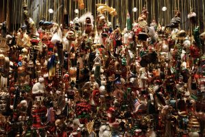 A dense collection of various Christmas ornaments hanging on strings with a dark background. The ornaments are diverse in shape and color, including figures of Santa Claus, animals, bells, and other festive decorations.
