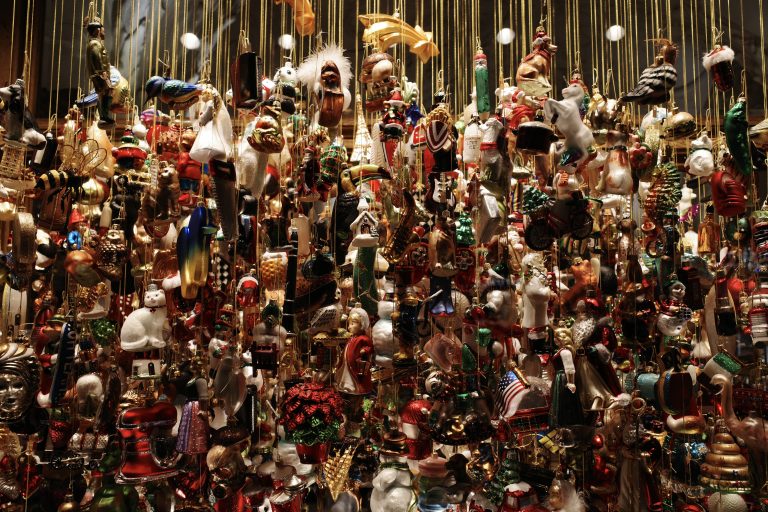 A dense collection of various Christmas ornaments hanging on strings with a dark background. The ornaments are diverse in shape and color, including figures of Santa Claus, animals, bells, and other festive decorations.