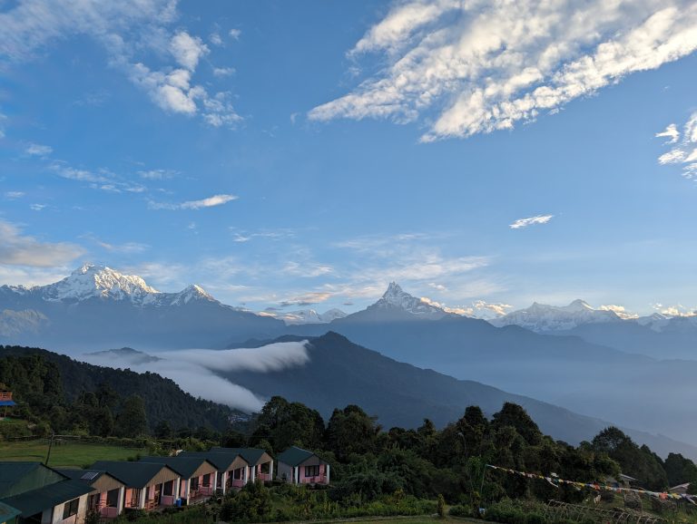 A scenic view of snow-capped Himalayan range mountain peaks from Australian Base Camp at sunrise with a layer of clouds below, a blue sky overhead, and a row of small structures in the foreground amidst green foliage.