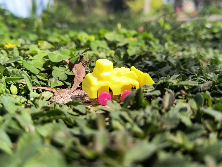 Close-up of a yellow toy car with pink wheels on green grass, with leaves and soil visible in the background.