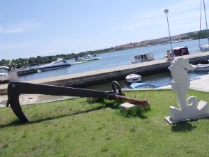 A large black anchor on display at a grassy seaside area with boats in the background and a white seahorse sculpture adjacent to the anchor