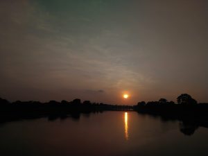 A serene sunset with the sun low in the sky reflecting on a calm river, against a backdrop of a hazy sky with scattered clouds and silhouetted trees on the horizon.