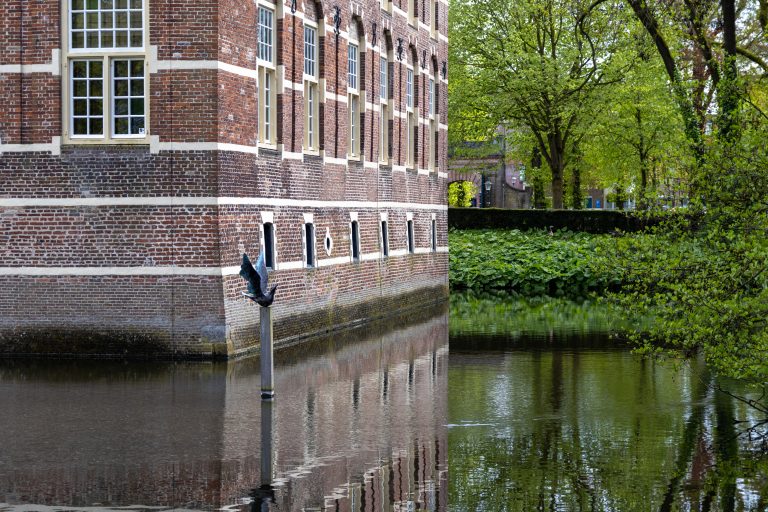 The corner of a castle, with moat around it with a sculpture of a flying bird in it, green trees and bushes.