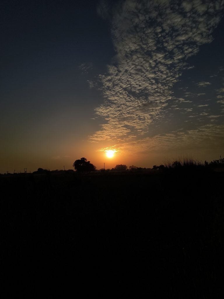 Sun setting over a rural landscape, with clouds scattered across the sky and silhouettes of trees and grass visible in the foreground.