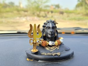 Close-up of a small idol of Lord Shiva placed on a dashboard, with a Trishul (trident) symbol in gold and beads around its neck.