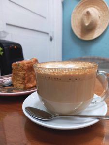 A close-up photo showing a glass cup of latte on a wooden table with a piece of layered cake in the background, against a backdrop of a blue door with a straw hat hanging on it.