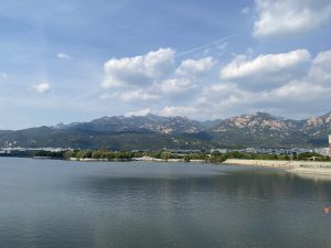 Calm water, green trees, blue sky, white clouds, mountains in the background
