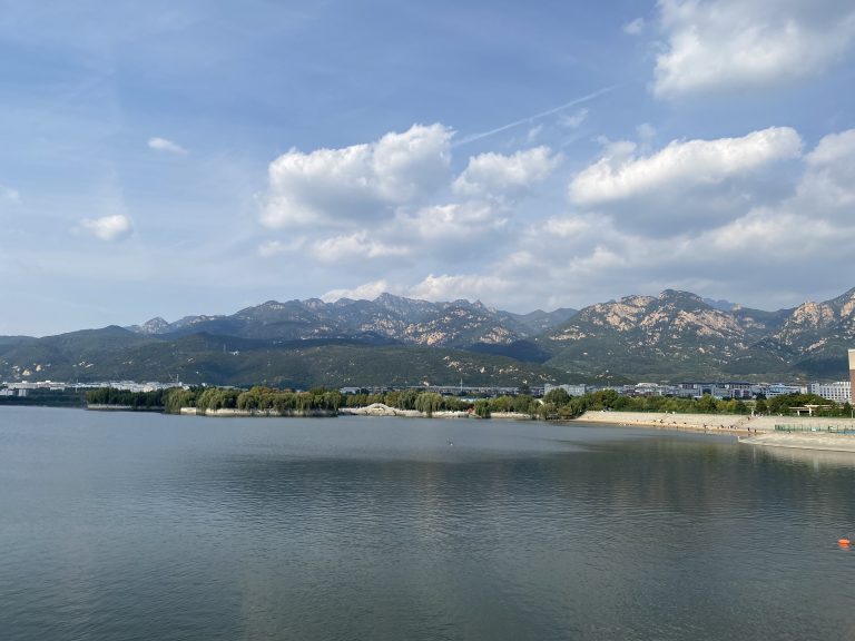 Calm water, green trees, blue sky, white clouds, mountains in the background