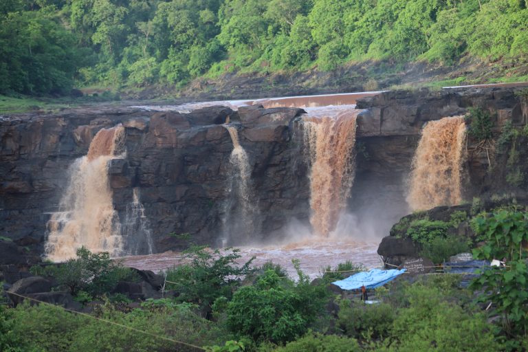 A scenic view of a waterfall with muddy brown water cascading down multiple rocky outcrops, surrounded by lush greenery. There are some blue tarpaulins and structures visible at the bottom of the image.