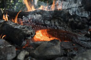 A close-up view of a wood fire with glowing embers and orange flames consuming charred logs, surrounded by grey ash and stones.
