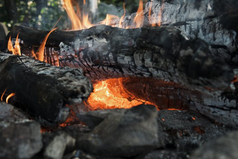 A close-up view of a wood fire with glowing embers and orange flames consuming charred logs, surrounded by grey ash and stones.