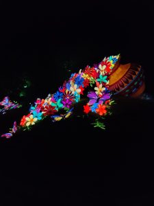 A colorful illuminated pot of a giant nautilus shell adorned with numerous bright flowers and butterflies, set against a dark night background.
