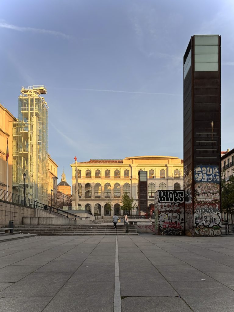 A person walking towards a Reina Sofía museum in Madrid, flanked by two modern structures with heavy graffiti on the side, under a clear blue sky with a contrail.