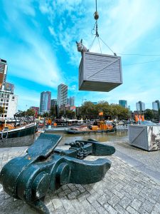 A statue in a port of rotterdam, a horse is sitting in a crate being lifted by a crane. Underneath the crate there is a big anchor lying on the pavement.