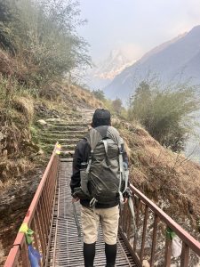 View larger photo: Photo Capture on the way to Annapurna Base Camp: trekkers with backpacks walking across a metal bridge on a mountain trail with a backdrop of snowy peaks partially obscured by mist.