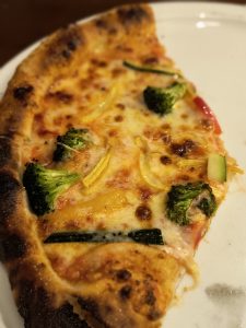 The image shows a close-up of a slice of pizza with melted cheese, tomato sauce, and various toppings such as broccoli, zucchini, and peppers.