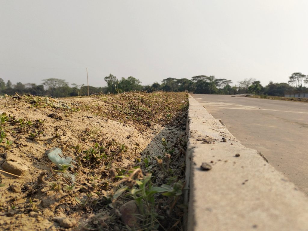 A close-up view of the edge of an asphalt road with a concrete curb, adjacent to a dusty, grassy area under a clear sky.