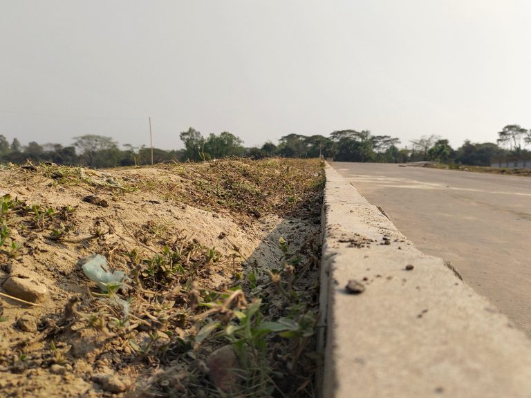 A close-up view of the edge of an asphalt road with a concrete curb, adjacent to a dusty, grassy area under a clear sky.