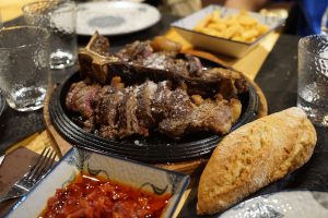 

A meal featuring a sliced beef steak on a skillet, sprinkled with salt, served with a side of fries in a metal basket and tomato-based sauce in a small dish, accompanied by a loaf of bread, with empty glasses and a patterned plate visible in the background.
