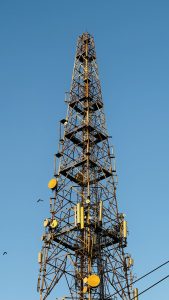 A tall communications tower with various antennas and dishes against a clear blue sky, with birds flying nearby.