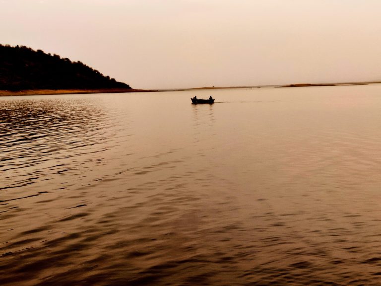 Two people row a boat in calm waters near a beach with hillside, at sunset under an orange-brown hue, possibly due to smog.