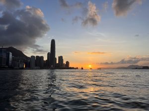 A sunset view over the water showcasing the silhouette of the hong kong skyline with skyscrapers against a colorful sky with scattered clouds.
