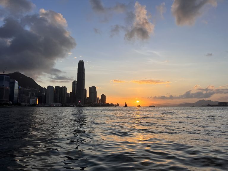 A sunset view over the water showcasing the silhouette of the hong kong skyline with skyscrapers against a colorful sky with scattered clouds.