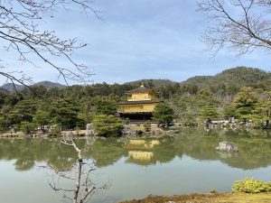 

A traditional golden Japanese pavilion reflected in a calm pond surrounded by pine trees and with a backdrop of forest-covered hills against a blue sky.