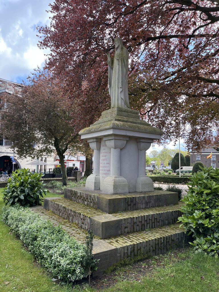 A statue of Christ under a tree with red leaves, between bushes and grass